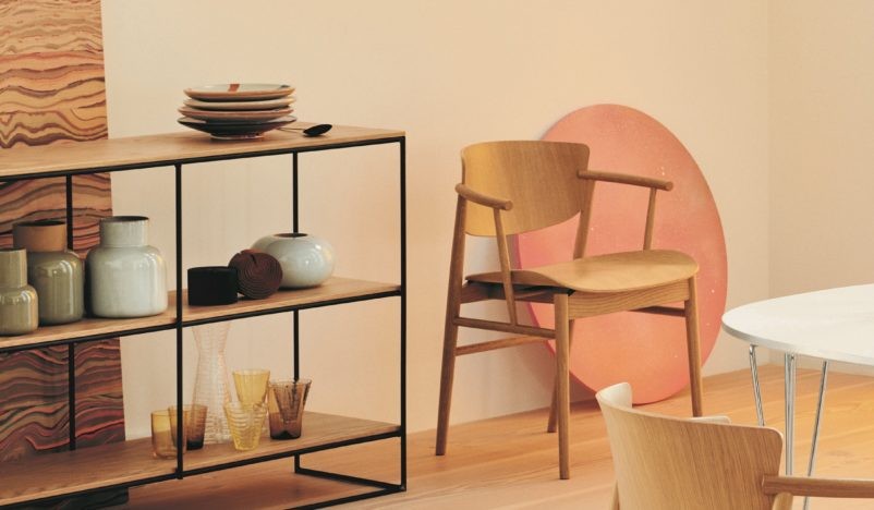 A modern dining room setting with a wooden chair, a black metal and wood shelving unit displaying ceramic vases and glassware, and a round dining table with wooden chairs. A large, pink circular art piece leans against the wall in the background.