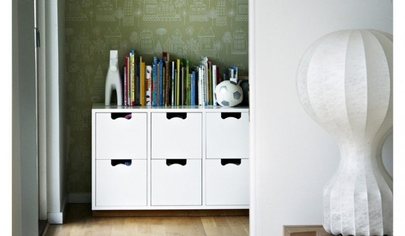 A cozy reading corner featuring a small white cabinet with four compartments, topped with an assortment of children's books, a decorative vase, and a soccer ball.