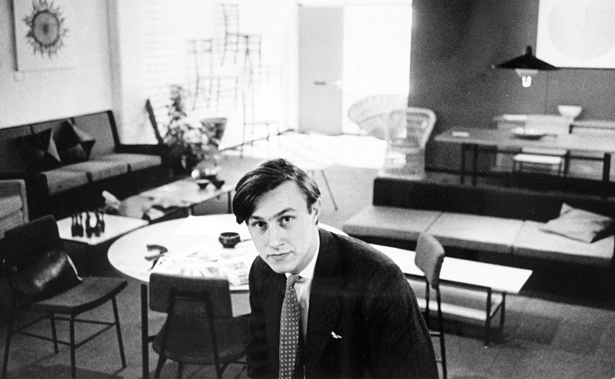 A black and white photograph of Terence Conran in a suit sitting in a stylish, mid-century modern living room with various furniture pieces and decor.