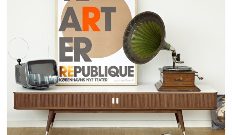 A vintage setup featuring a mid-century modern wooden sideboard, an old-fashioned television with an antenna, a gramophone, and a large art poster leaning against the wall.