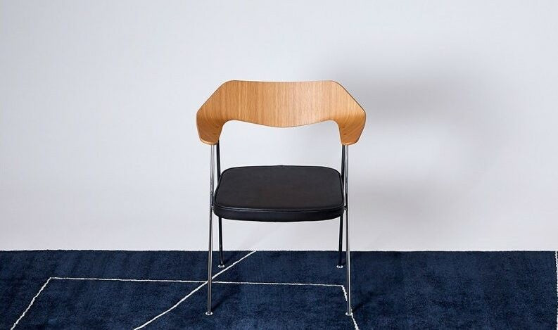 A wooden chair with a black seat cushion on a dark blue rug with white geometric lines.