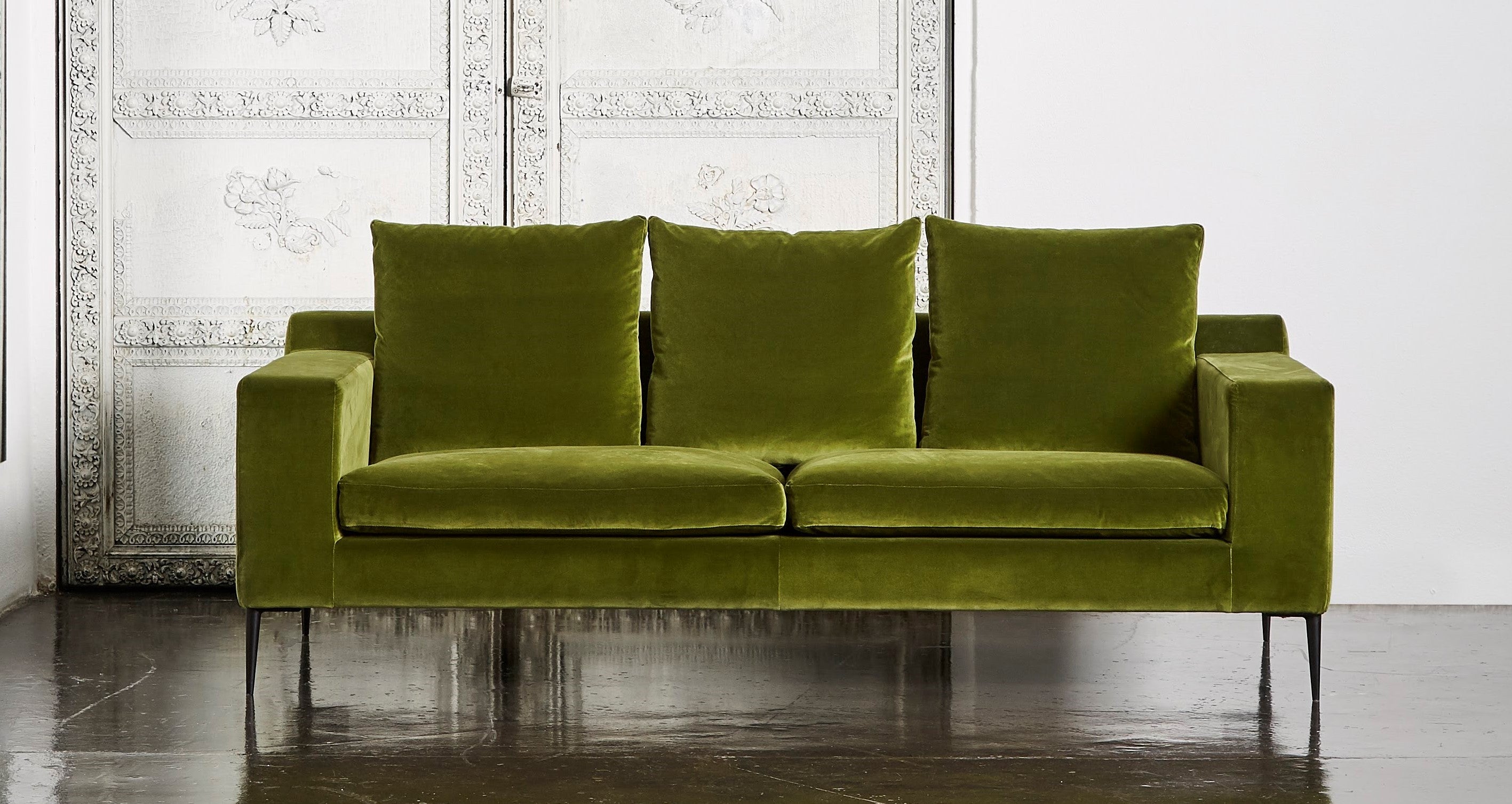 A luxurious olive green velvet sofa set against an ornate white carved door backdrop. The sofa features a deep, plush seating area with three back cushions enhancing its inviting appearance.