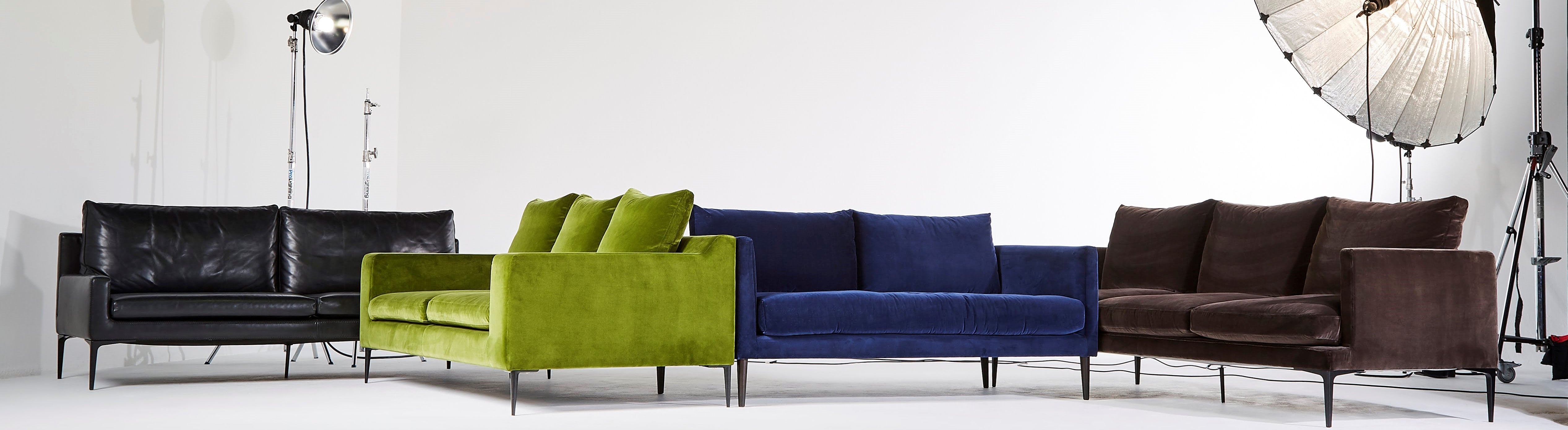 This image displays a collection of five elegant sofas, each showcasing distinct styles and colors, set against a minimalist white studio background. From left to right: a sleek black leather sofa, a vibrant lime green velvet sofa with matching back cushions, a luxurious deep blue velvet sofa, a classic dark brown leather sofa, and a retro-style chocolate brown velvet sofa with plush cushions.