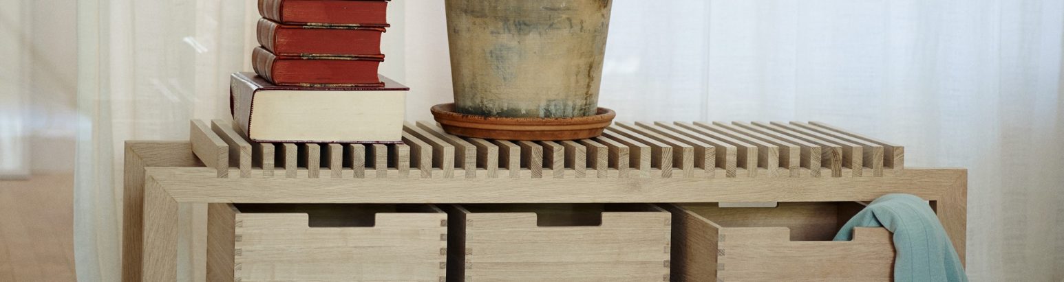 A close-up of a wooden storage unit with an open slatted top, featuring three drawers, a stack of books, and a potted plant.