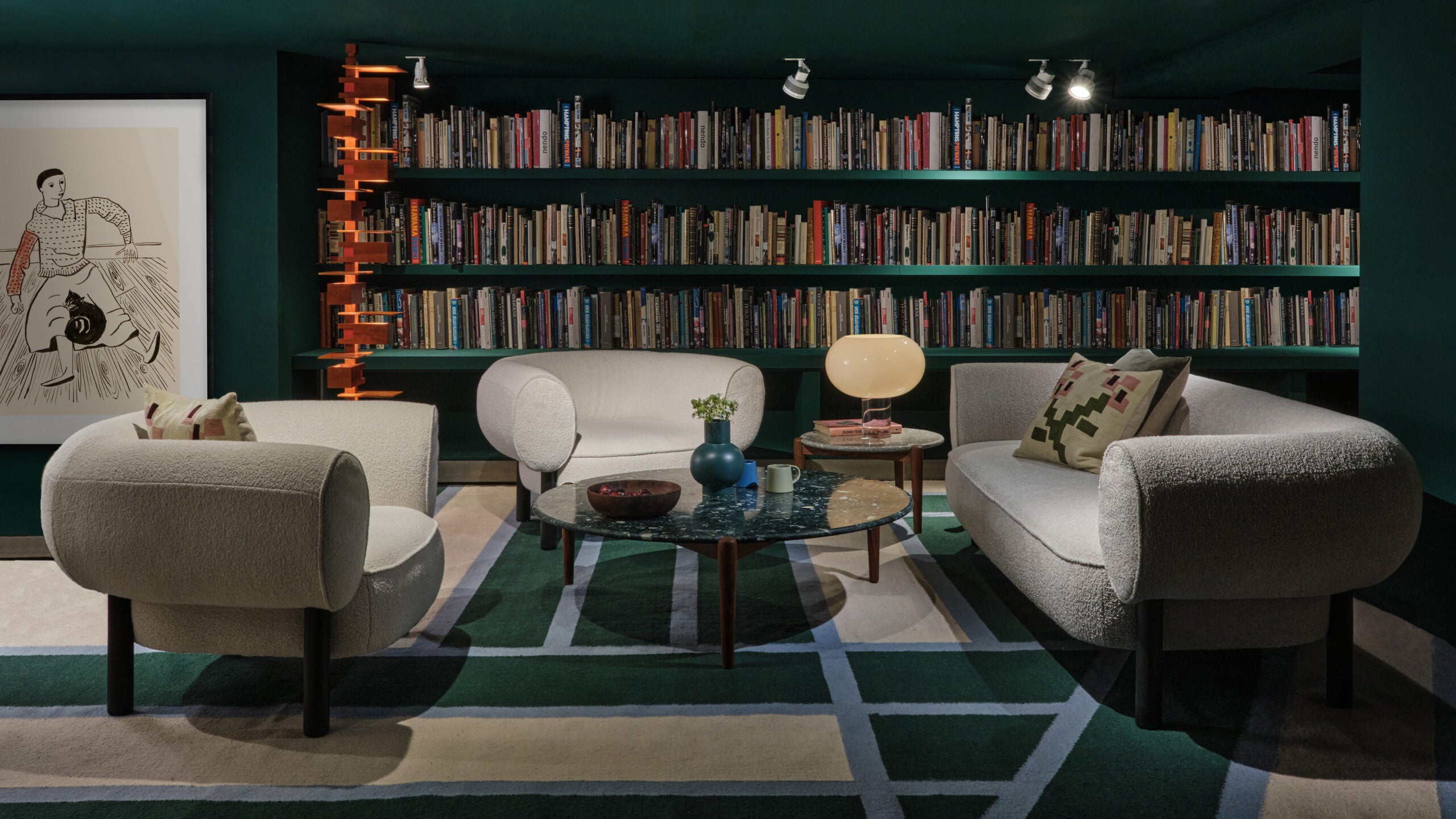 A cozy living room setting featuring plush white sofas and armchairs, a circular marble coffee table, and a bookshelf filled with books against a dark green wall. An abstract art piece and modern lighting add to the sophisticated ambiance.