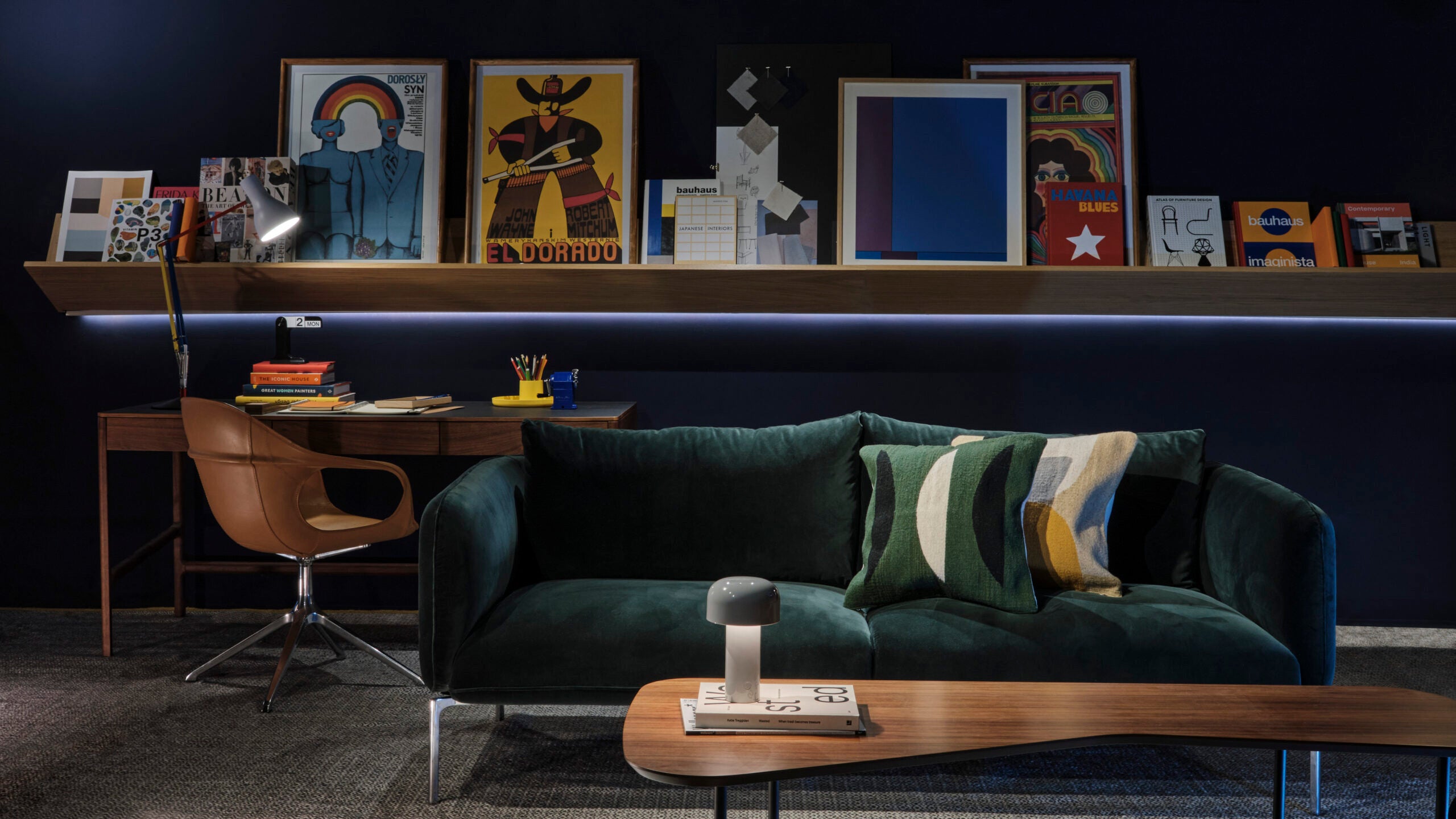 A sophisticated home office setup with a dark theme. The room features a plush emerald green velvet sofa adorned with striped cushions in shades of green, yellow, and gray. A sleek wooden desk with a stylish leather swivel chair is set against a navy blue wall lined with an illuminated shelf displaying an assortment of colorful art and design books.