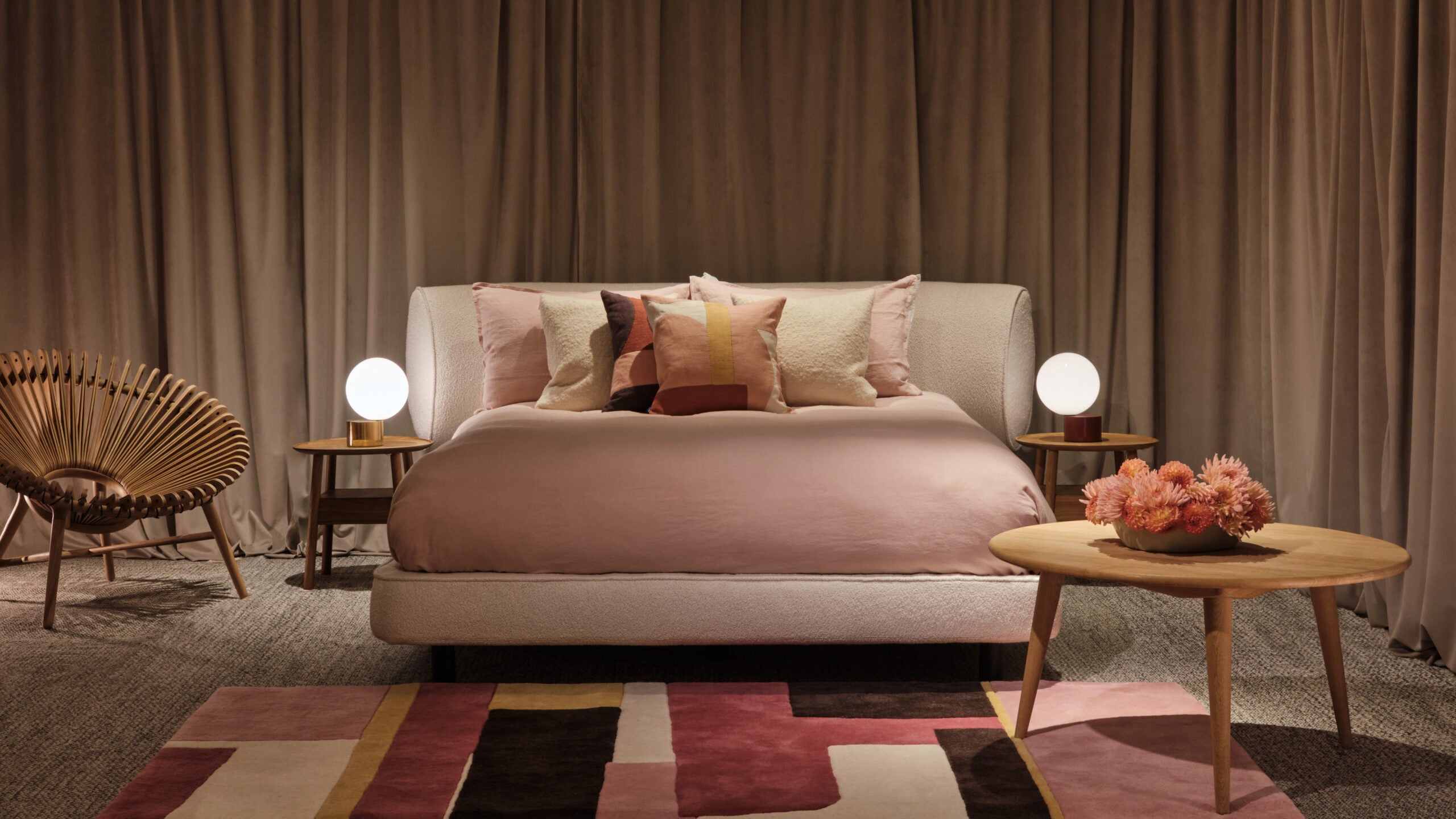 A stylish bedroom features a plush bed with pink bedding, decorative pillows, modern side tables with globe lamps, and a colorful geometric rug.