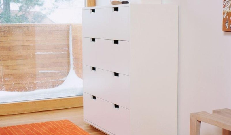 A modern white dresser with four drawers is placed in a bright room with wooden floors and a large window, overlooking a snowy outdoor scene.