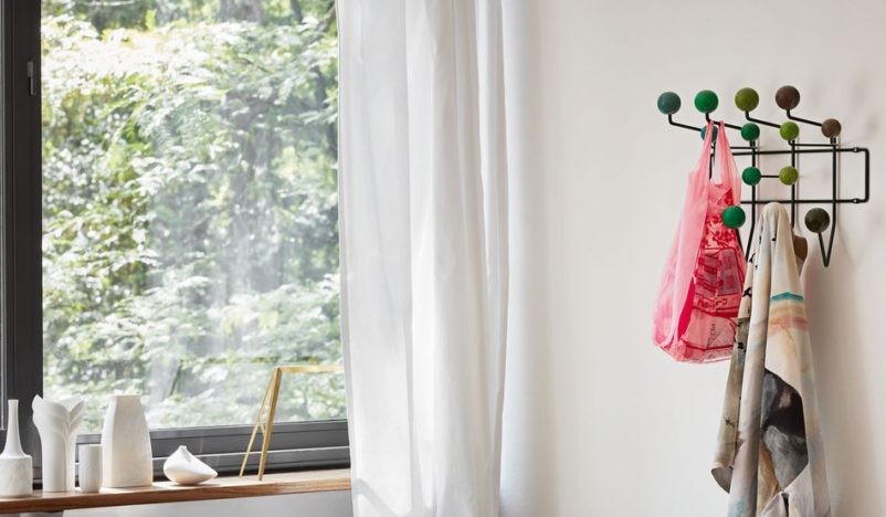 A brightly lit room with a window on the left, featuring white curtains and a wooden windowsill adorned with minimalist ceramic vases. To the right, a wall-mounted coat rack with colorful round hooks holds a pink bag and a patterned scarf.