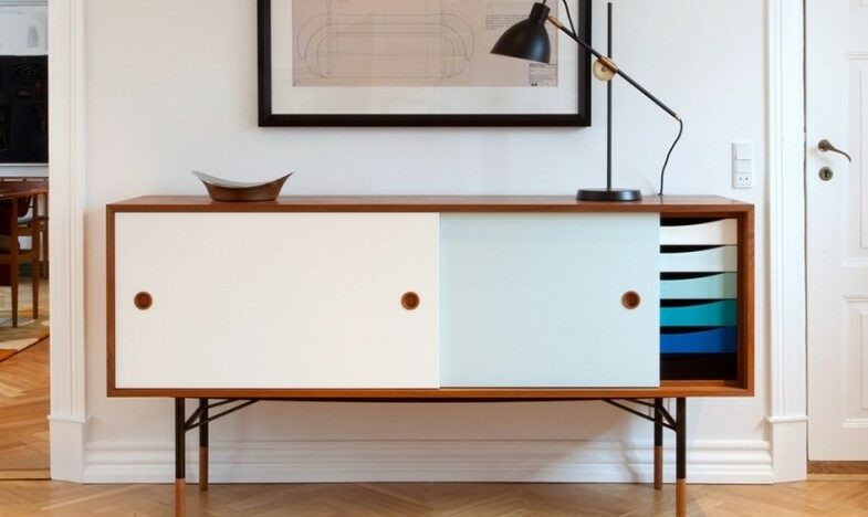 A mid-century modern sideboard with a white and light blue sliding door, showcasing a minimalist design. The sideboard is complemented by a black desk lamp, a decorative bowl, and framed artwork above it.