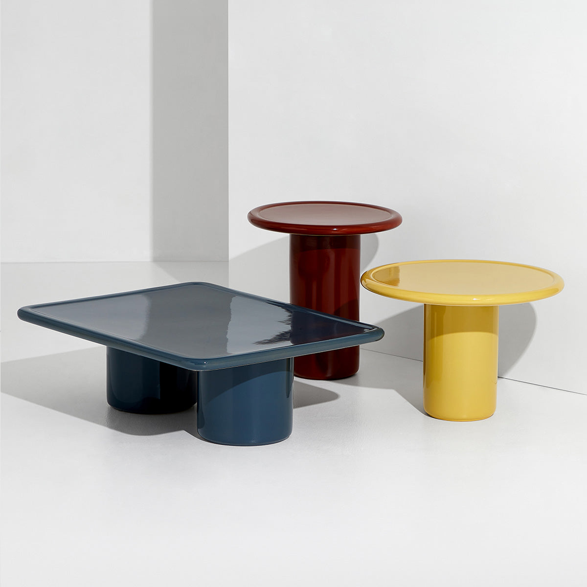 A set of three Mag coffee tables designed by Daniel Schofield in blue, red, and yellow finishes, featuring round and square tops, displayed against a white background.