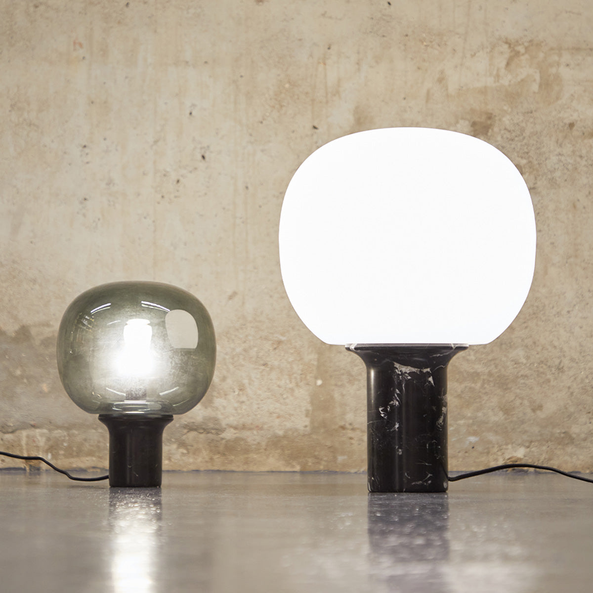 Two Flo modern table lamps with black bases are placed on a concrete floor against a textured wall, one with a large white shade and the other with a smaller smoky gray shade.