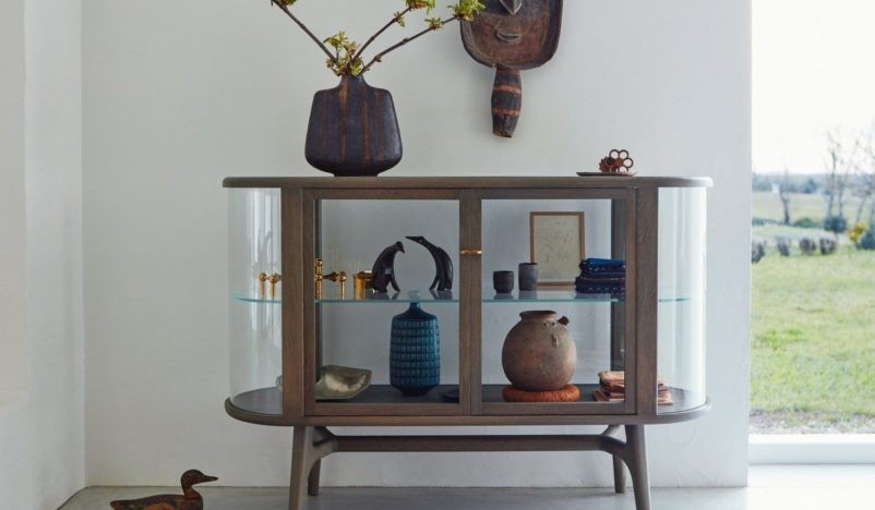 A stylish glass display cabinet with wooden frame showcasing various decorative items, topped with a vase holding branches, and an African mask mounted on the wall above it, set in a bright room with a view of a grassy landscape outside the window.