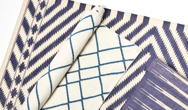 A collection of rugs featuring different geometric patterns in blue and white. One rug has a zigzag design, another displays a grid pattern, and a third has linear stripes, all arranged in an overlapping fashion.