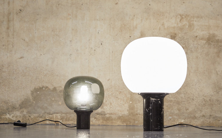 Two distinctive lamps placed against a rough concrete wall, illustrating a study in contrasts. On the left, a transparent glass lamp reveals its inner bulb and filament. To the right, a lamp with a solid black base supports a luminous white globe, emitting a bright, diffused light. T