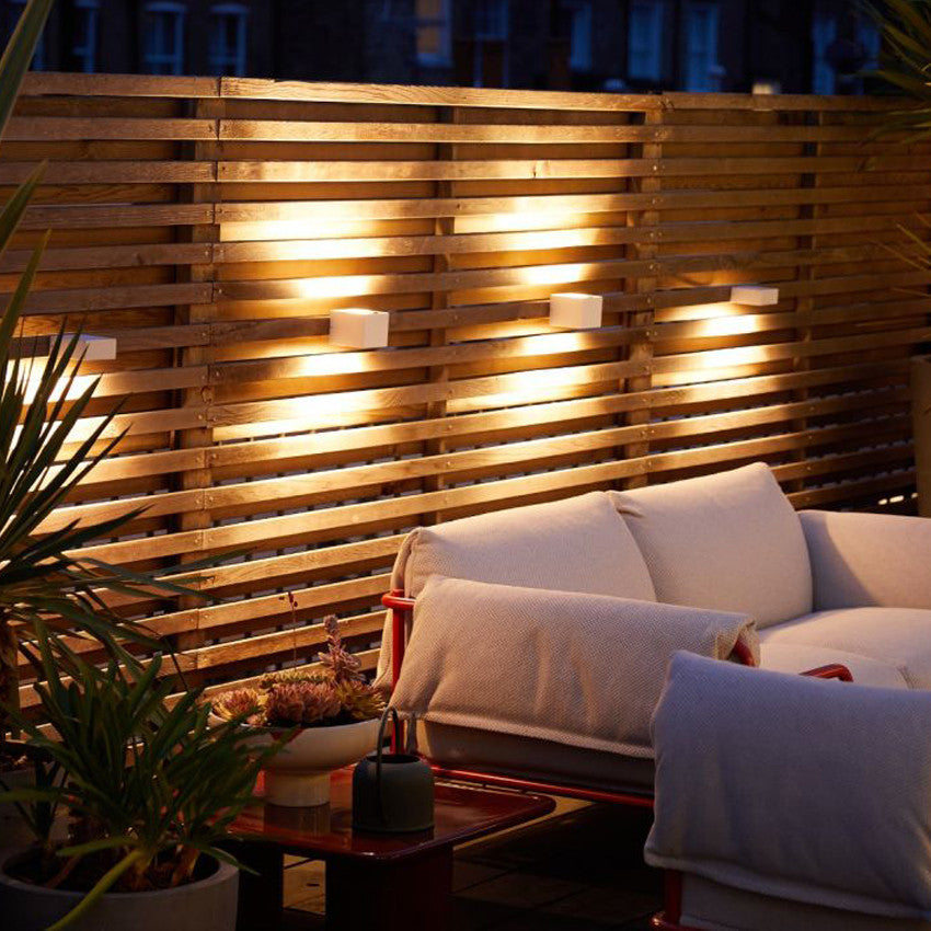 A cozy outdoor seating area at night features a wooden slatted wall illuminated by warm wall lights, with a Fold Outdoor 2-Seater sofa and potted plants.