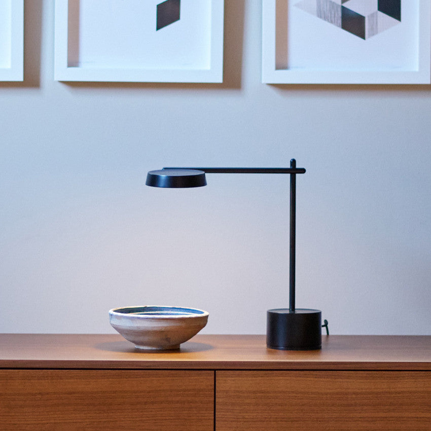A modern black Roto desk lamp placed on a wooden sideboard, next to a ceramic bowl, with framed geometric artwork on the wall behind.