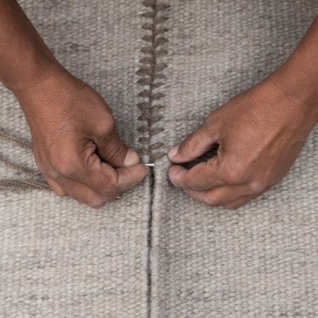 Hands of a craftsman stitching together pieces of a grey wool rug with intricate detail.