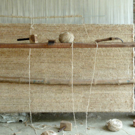 A rustic weaving loom setup featuring natural fiber yarns hanging vertically from a wooden beam. The loom, crafted from weathered wood, is partially covered with woven fabric displaying a coarse texture.