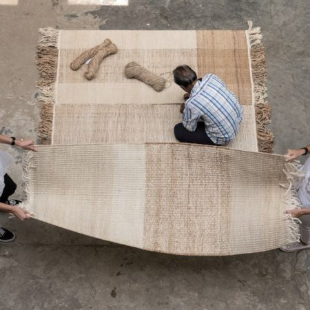 Two people hold a large woven rug while a third person, seated, works on it with bundles of yarn beside them. The rug has a patchwork design in shades of beige and brown, with fringed edges.