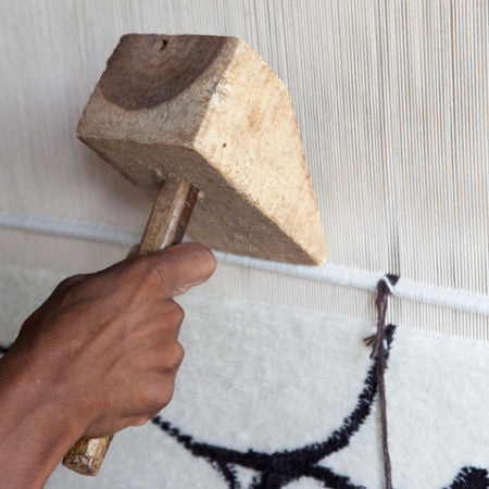 Close-up of a hand wielding a traditional wooden mallet, striking a loom to adjust or secure the weave of a white and black patterned rug.