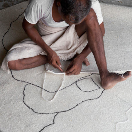 A craftsman is sitting cross-legged on the floor, weaving a white rug with black abstract lines. He is wearing a white shirt and a beige cloth wrapped around his waist, meticulously working with the thread and loom.