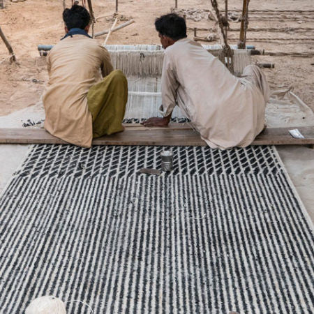 Two artisans in traditional clothing are weaving a striped rug on a loom outdoors, highlighting the craftsmanship involved in creating handmade textiles.