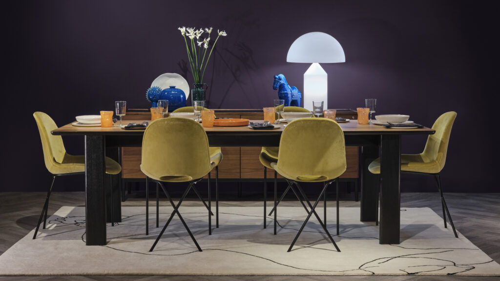 A modern dining room setup featuring a rectangular wooden table surrounded by six mustard yellow chairs with black metal legs. The table is set with various dishes and glassware, while a white lamp, blue horse statue, and vases with white flowers adorn the sideboard against a dark purple wall in the background.