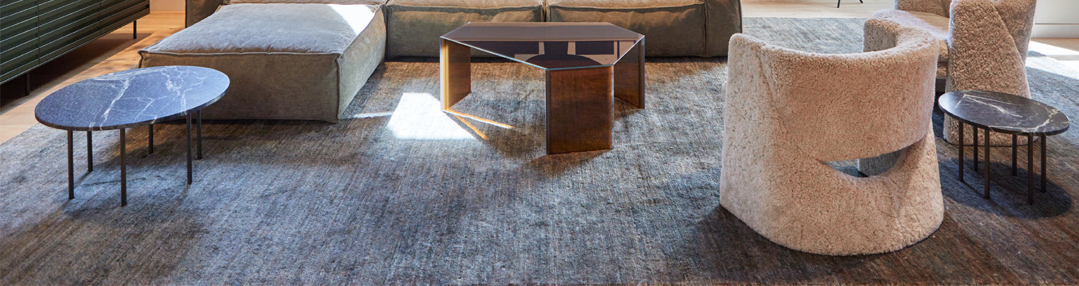 A close-up of a living room featuring a plush grey rug, a large ottoman, two modern side tables with marble tops, a geometric coffee table, and two cozy, curved armchairs.