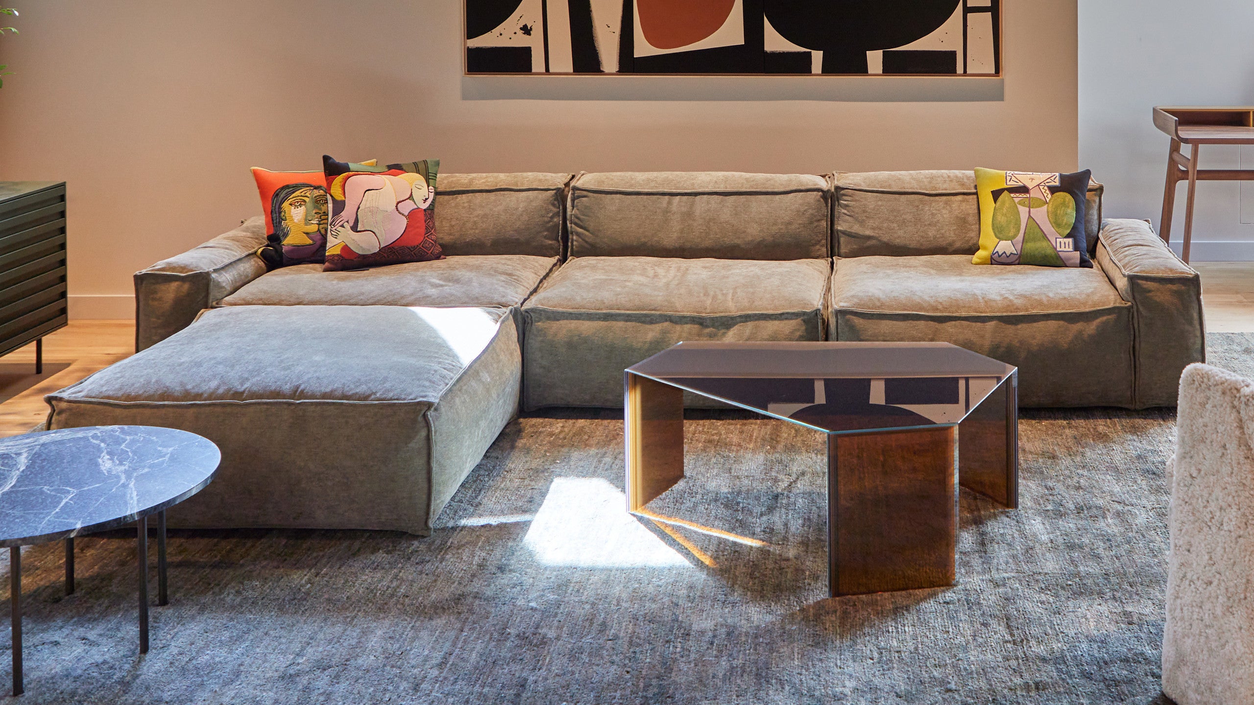 A cozy living room featuring a large, cushioned sectional sofa in a muted tone. The sofa is decorated with colorful throw pillows, including one with a modern art-inspired design and another with a playful character motif.