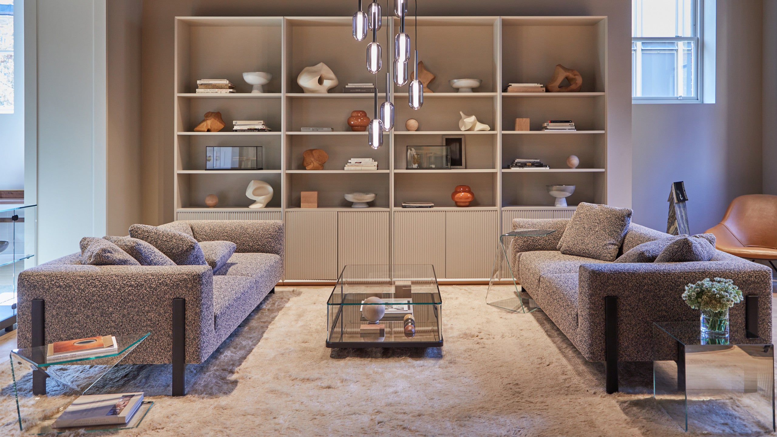 A modern living room featuring two gray sofas facing each other with a glass coffee table in between, set on a plush beige rug. Behind the sofas is a large bookshelf filled with books and various decorative items, and a cluster of pendant lights hangs from the ceiling.